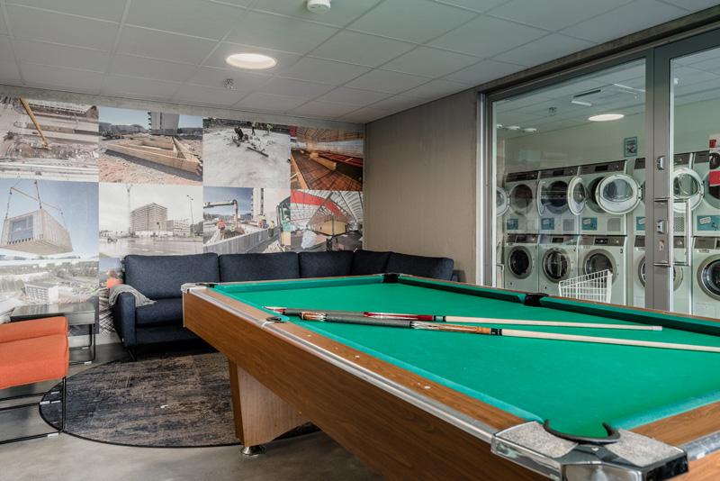 Pool table in the front of the picture. Behind that is a large couch. On the side wall is a glass wall through which the laundry rooms washing machines can be seen.